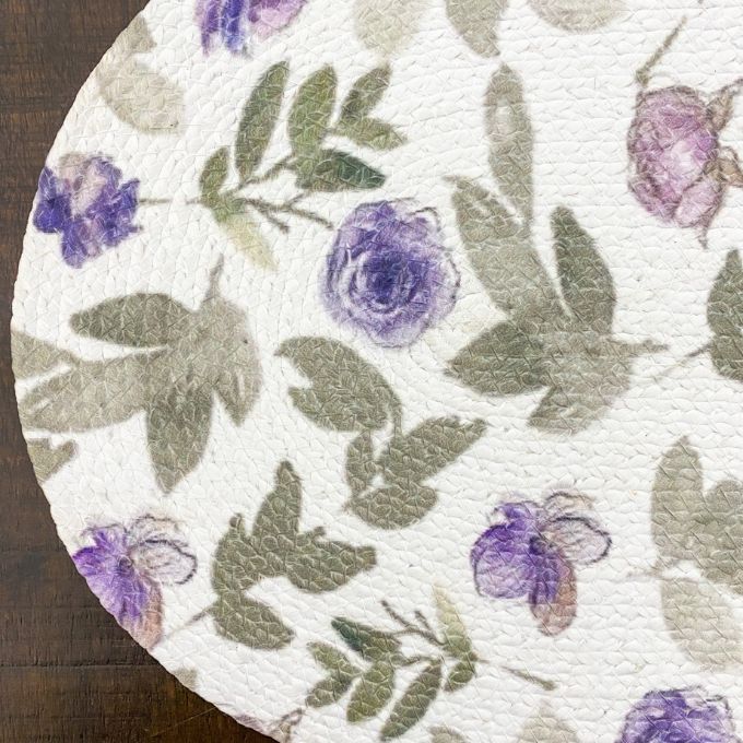 Round white mat with purple floral and green leaf pattern on a wooden surface. Violet Meadow Table Linen Collection - Three Styles available at Quilted Cabin Home Decor.