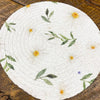 Round woven mat with floral patterns on a wooden surface. Daisy Blossom Braided Table Linens - Three Styles available at Quilted Cabin Home Decor.