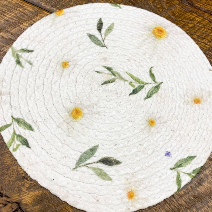 Round woven mat with floral patterns on a wooden surface. Daisy Blossom Braided Table Linens - Three Styles available at Quilted Cabin Home Decor.