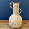 Textured ceramic vase with handles on a wooden surface against a blue wall. Rustic Metal Vases - Two Colours available at Quilted Cabin Home Decor.