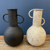 Two textured vases, one black and one white on a wooden surface with a blue background. Rustic Metal Vases - Two Colours available at Quilted Cabin Home Decor.