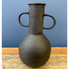 Black textured vase with handles on a wooden surface and blue background. Rustic Metal Vases - Two Colours available at Quilted Cabin Home Decor.