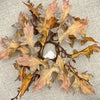 A circular autumn leaf wreath made of dried oak leaves in shades of brown, tan, and muted pink, attached to a dark brown branch base, laid flat on a textured beige fabric background. Autumn leaf candle ring available at Quilted Cabin in Airdrie