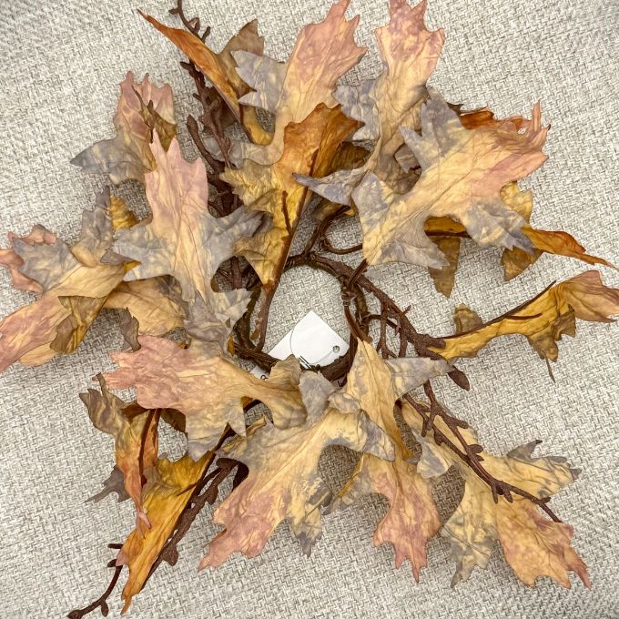 A circular autumn leaf wreath made of dried oak leaves in shades of brown, tan, and muted pink, attached to a dark brown branch base, laid flat on a textured beige fabric background. Autumn leaf candle ring available at Quilted Cabin in Airdrie