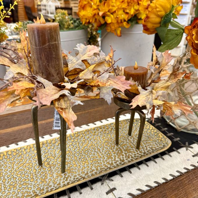 An autumn-themed centerpiece arrangement with dried leaves, two small candles, and a large pillar candle on a patterned rectangular tray in a store setting. All items available at Quilted Cabin in Airdrie