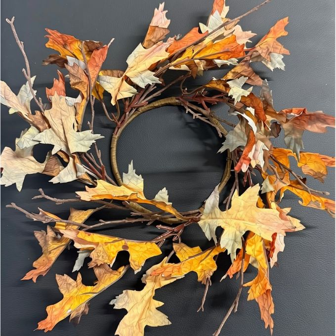 A circular autumn leaf wreath made of dried oak leaves in shades of brown, tan, and muted pink, attached to a dark brown branch base, laid flat on a textured beige fabric background. Autumn leaf candle ring available at Quilted Cabin in Airdrie