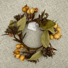 A decorative artificial wreath with olive-green leaves, brown branches, and small round tan berries, laid flat on a textured light gray fabric background. A white paper tag is attached near the center. Available at Quilted Cabin in Airdrie