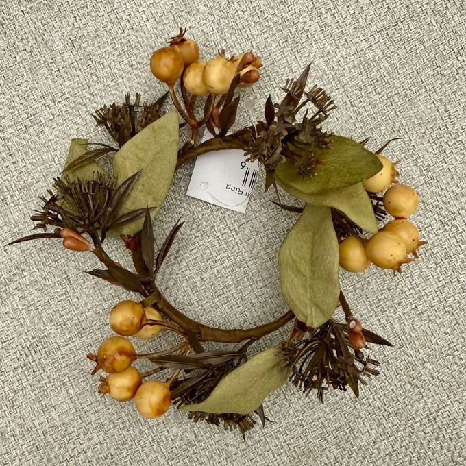 A decorative artificial wreath with olive-green leaves, brown branches, and small round tan berries, laid flat on a textured light gray fabric background. A white paper tag is attached near the center. Available at Quilted Cabin in Airdrie