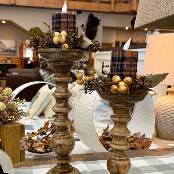 Two rustic wooden candlestick holders with plaid pillar candles, decorated with faux berries and leaves, displayed on a table in a cozy home setting.  Florals available at Quilted Cabin in Airdrie