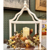 A white distressed wooden lantern centerpiece with a cream pillar candle inside, surrounded by autumn foliage, dried leaves, and small yellow berries, displayed against a dark green pegboard wall.  All products available at Quilted Cabin in Airdrie