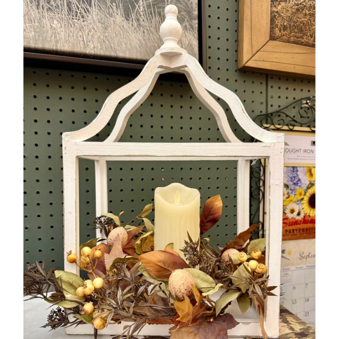 A white distressed wooden lantern centerpiece with a cream pillar candle inside, surrounded by autumn foliage, dried leaves, and small yellow berries, displayed against a dark green pegboard wall.  All products available at Quilted Cabin in Airdrie
