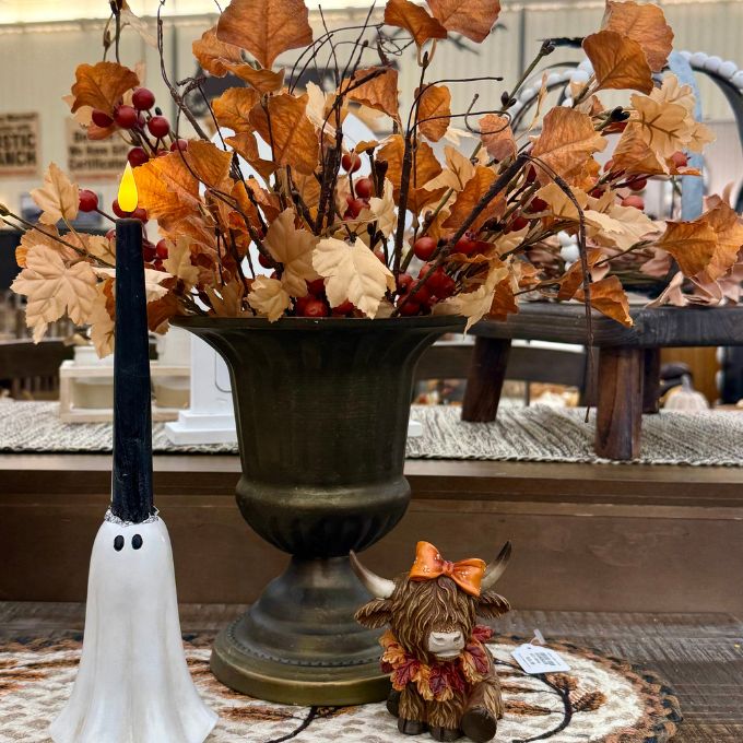 An autumn-themed tabletop arrangement featuring a large bronze-colored urn filled with faux maple leaves in shades of brown and beige, accented with small red berries. To the left is a black taper candle shaped like a white ghost with a glowing flame. All decor items and florals available at Quilted Cabin in Airdrie