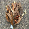 A decorative artificial autumn wreath or floral arrangement made of dried brown and tan leaves, wheat-like stalks, pinecones, and small round brown berries, photographed on a gravel surface.  Available at Quilted Cabin in Airdrie