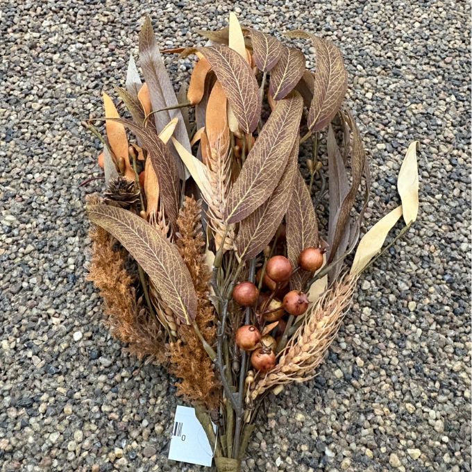 A decorative artificial autumn wreath or floral arrangement made of dried brown and tan leaves, wheat-like stalks, pinecones, and small round brown berries, photographed on a gravel surface.  Available at Quilted Cabin in Airdrie