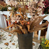 Autumn floral arrangement in a ribbed dark gray ceramic pot, featuring beige flowers, brown leaves, and small orange berries, displayed on a patterned tablecloth in a shop setting.  All items available at Quilted Home in Airdrie