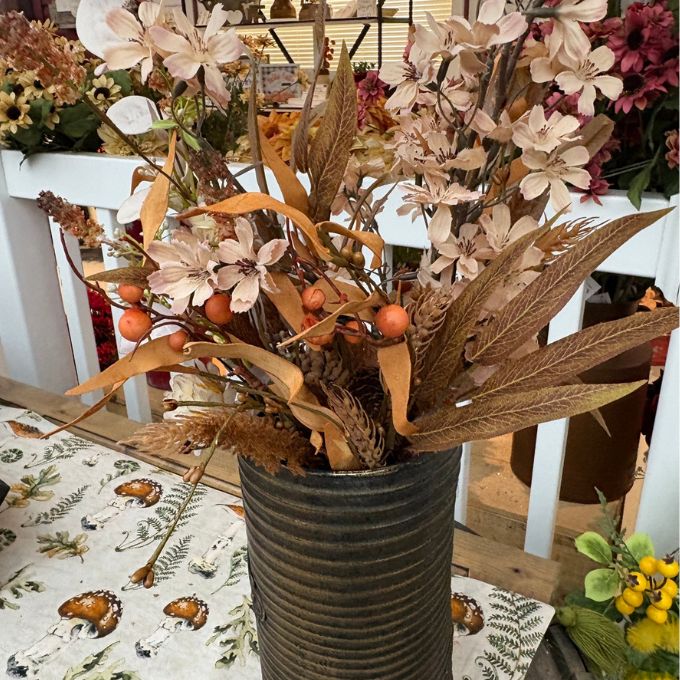 Autumn floral arrangement in a ribbed dark gray ceramic pot, featuring beige flowers, brown leaves, and small orange berries, displayed on a patterned tablecloth in a shop setting.  All items available at Quilted Home in Airdrie