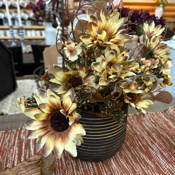 A decorative artificial floral arrangement with large sunflower-like blooms in cream, tan, and brown tones, set in a dark ribbed vase on a patterned table runner.  Available at Quilted Cabin in Airdrie