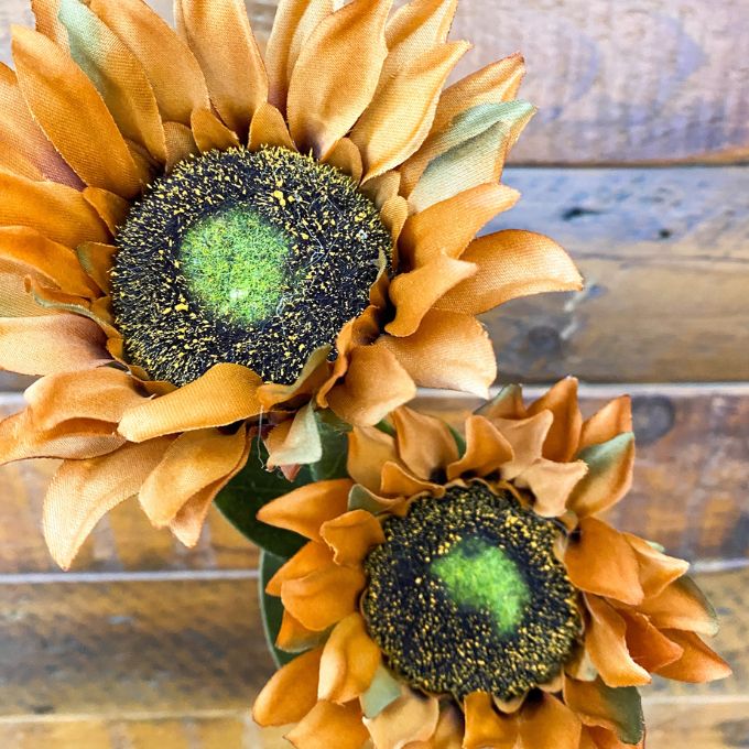 Two artificial sunflowers with orange petals and green centers on a wooden surface. Sunflower Sprays - Two Colours available at Quilted Cabin Home Decor.