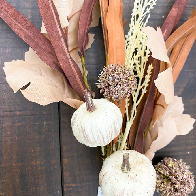 Decorative arrangement with white pumpkins, dried flowers, and ribbons on a dark wooden surface. Pumpkin and Oak Leaf Candle Ring and Floral Spray available at Quilted Cabin Home Decor.