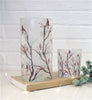 Two frosted glass candle holders with a bird and branch pattern, displayed on a book against a white brick wall background available at Quilted Cabin in Airdrie