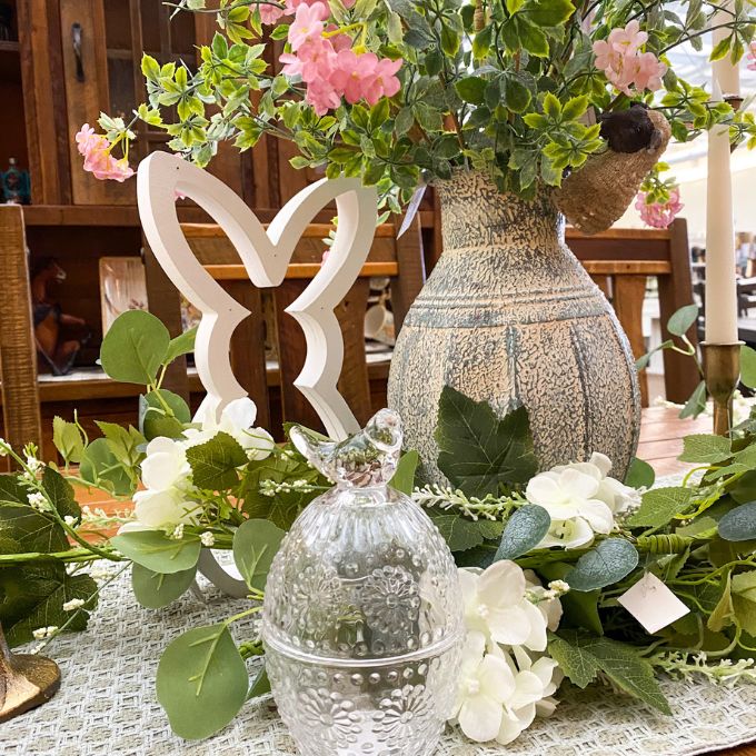 Decorative table setting with flowers, a vase, and a glass container on a tablecloth. Clear Glass Egg Shaped Dish with Bird Finial available at Quilted Cabin Home Decor.