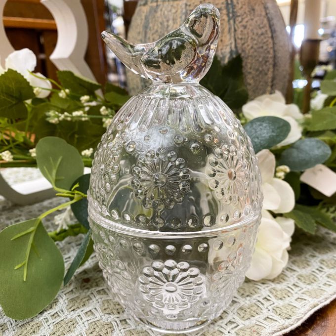 Decorative glass egg shaped jar with floral patterns on a table with flowers and greenery.  Clear Glass Egg Shaped Dish with Bird Finial available at Quilted Cabin Home Decor.