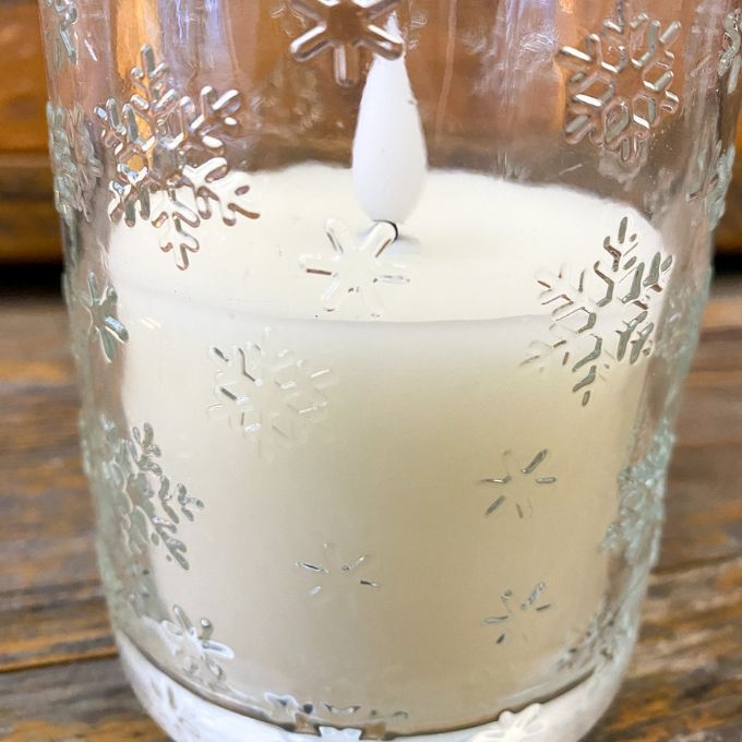 White candle in a glass holder with snowflake designs on a wooden surface. Snowflake Glass LED Timer Candle available at Quilted Cabin Home Decor.