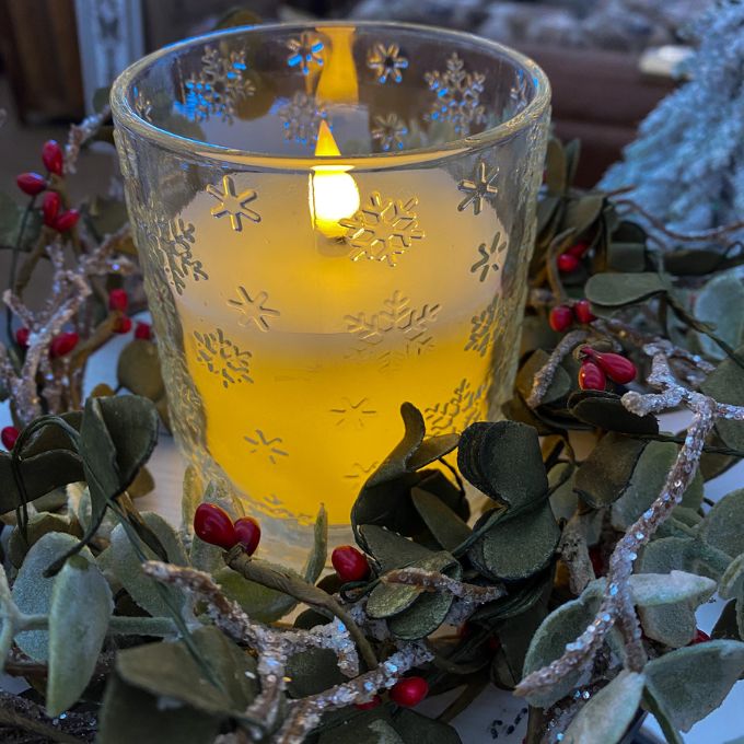 Candle in a decorative glass holder with snowflake patterns, surrounded by greenery and red berries. Snowflake Glass LED Timer Candle available at Quilted Cabin Home Decor.