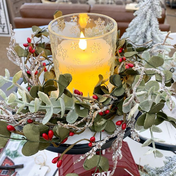 Decorative wreath with a lit candle surrounded by greenery and red berries on a table. Snowflake Glass LED Timer Candle available at Quilted Cabin Home Decor.