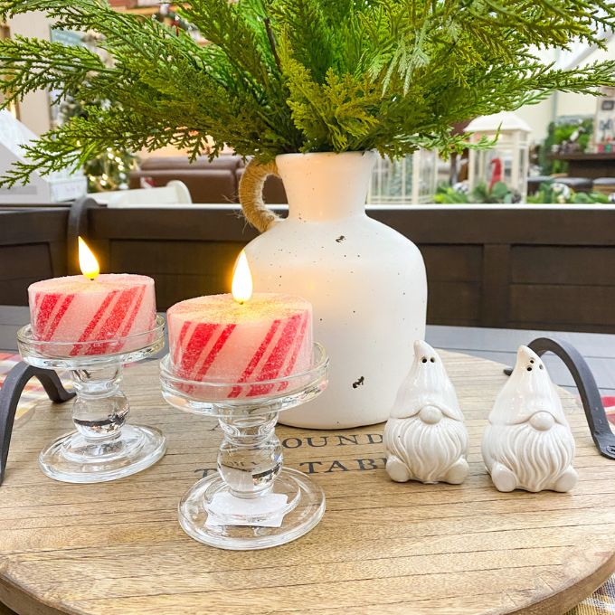 Two lit candles in glass holders with a vase and two small gnomes on a wooden table.  Simple Glass Candle Holder available at Quilted Cabin Home Decor.