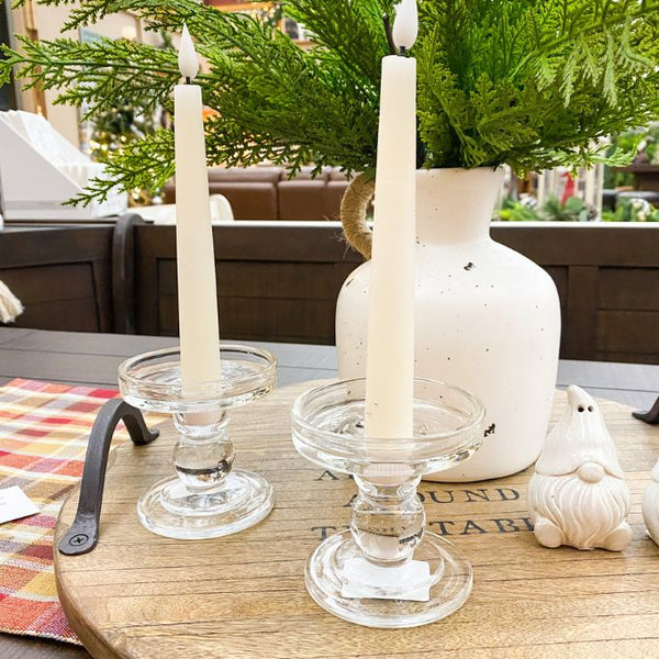 Two white candles in glass candle holders on a wooden table with a vase and greenery in the background. Simple Glass Candle Holder available at Quilted Cabin Home Decor.