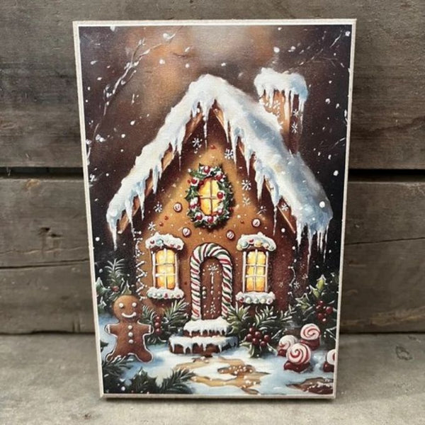 A vertical rectangular Christmas art print or card featuring a snow-covered gingerbread house with glowing windows, candy canes, a wreath, and a gingerbread man, displayed against a rustic wooden plank background available at Quilted Cabin in Airdrie