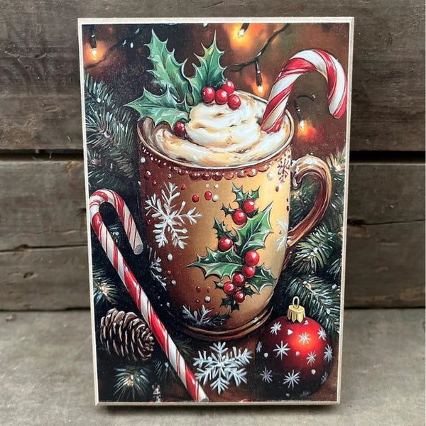 A rectangular Christmas-themed print or card leaning against a rustic wooden surface, showing a decorated brown mug filled with whipped cream and a candy cane, surrounded by holly, pine branches, candy canes, ornaments, and warm string lights.  Available at Quilted Cabin in Airdrie