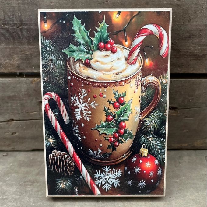 A rectangular Christmas-themed print or card leaning against a rustic wooden surface, showing a decorated brown mug filled with whipped cream and a candy cane, surrounded by holly, pine branches, candy canes, ornaments, and warm string lights.  Available at Quilted Cabin in Airdrie