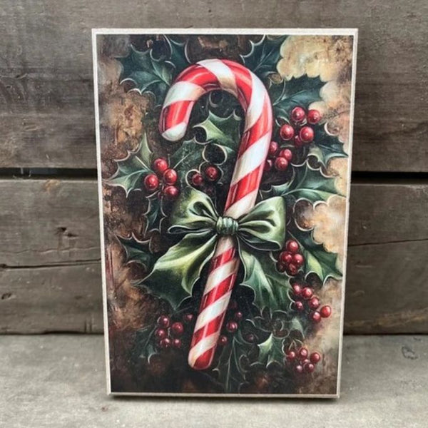 A rectangular Christmas art print or card featuring a large red-and-white striped candy cane surrounded by holly leaves and red berries, photographed leaning against a rustic wooden surface. Available at Quilted Cabin in Airdrie