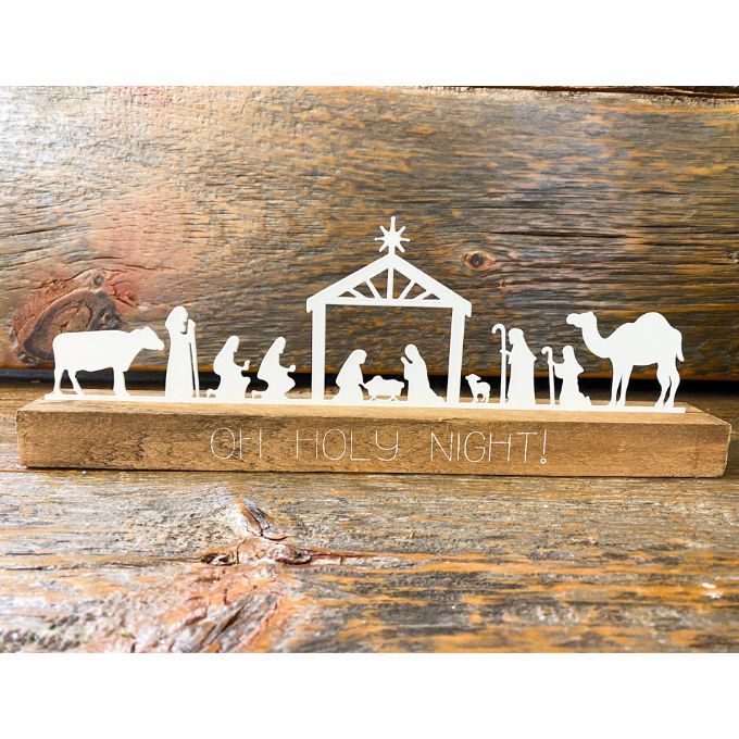 A white metal nativity scene with all characters in a row sits on a brown distressed board. The words on the board say Oh Holy Night. It is available at Quilted Cabin Home Decor.