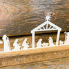 A white metal nativity scene with all characters in a row sits on a brown distressed board. The words on the board say Oh Holy Night. It is available at Quilted Cabin Home Decor.