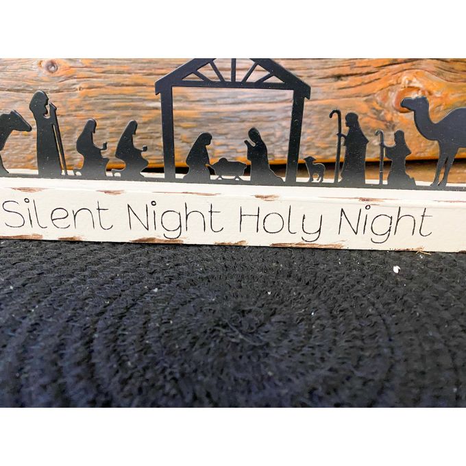 A black metal nativity scene with all characters in a row sits on a white distressed board. The words on the board say Silent Night Holy Night. It is available at Quilted Cabin Home Decor.