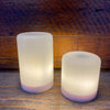 Two cylindrical LED lights on a wooden surface with a wooden background.  Illusion Pillar LED Candles - Two Sizes and Colours available at Quilted Cabin Home Decor.