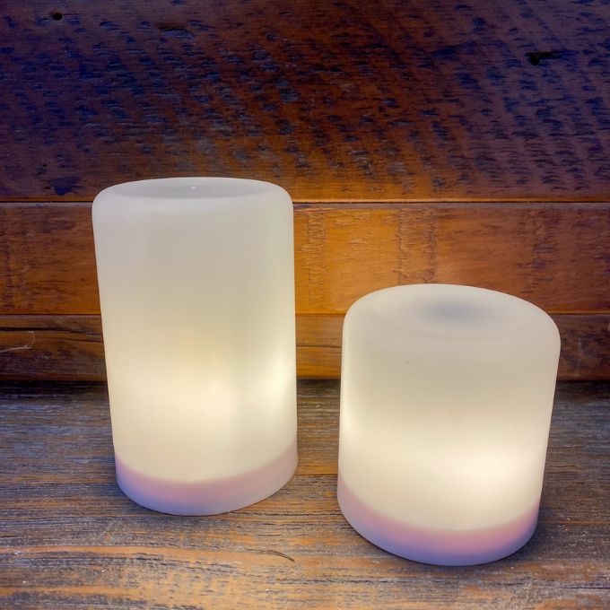Two cylindrical LED lights on a wooden surface with a wooden background.  Illusion Pillar LED Candles - Two Sizes and Colours available at Quilted Cabin Home Decor.