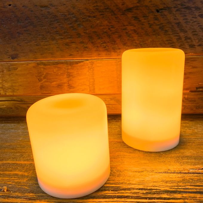Two glowing cylindrical LED pillars  on a wooden surface.  Illusion Pillar LED Candles - Two Sizes and Colours available at Quilted Cabin Home Decor.