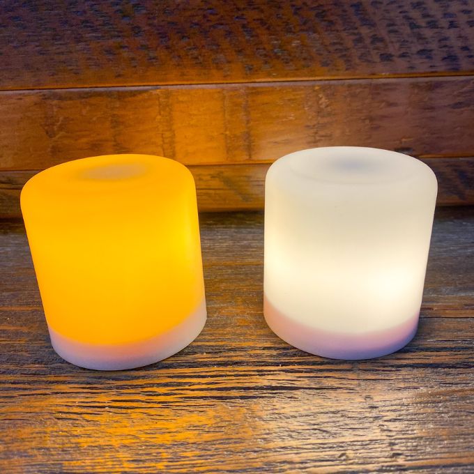 Two cylindrical LED lights, one orange and one white, on a wooden surface.  Illusion Pillar LED Candles - Two Sizes and Colours available at Quilted Cabin Home Decor.