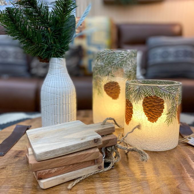 Decorative candles with pinecones and branches on a wooden table, surrounded by a cozy living room setting.  Illusion Pillar LED Candles - Two Sizes and Colours available at Quilted Cabin Home Decor.