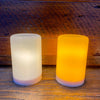 Two cylindrical LED lights, one white and one orange, on a wooden surface.  Illusion Pillar LED Candles - Two Sizes and Colours available at Quilted Cabin Home Decor.