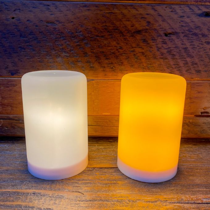 Two cylindrical LED lights, one white and one orange, on a wooden surface.  Illusion Pillar LED Candles - Two Sizes and Colours available at Quilted Cabin Home Decor.