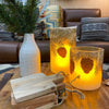Decorative lanterns with pine cone design on a wooden table in a cozy living room.  Illusion Pillar LED Candles - Two Sizes and Colours available at Quilted Cabin Home Decor.