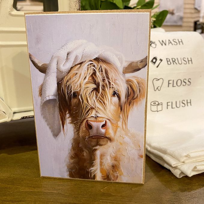Card with a highland cow wearing a towel on its head, placed on a surface with a towel and text in the background. Just Washed Block Art available at Quilted Cabin Home Decor.