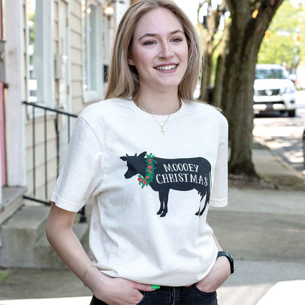 The Mooey Christmas T-shirt is an off white tshirt with a print of a black cow with a red and green wreath on its  neck and Mooey Christmas is printed on it. Available at Quilted Cabin Home Decor.