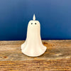 A small white ghost-shaped candle with black eyes sits on a rustic wooden surface against a dark blue background. Ghost candle available at Quilted Cabin in Airdrie