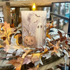 A lit cylindrical Halloween candle with a cream-colored surface printed with a ghost, pumpkin, moon, and bats, styled in a rustic autumn wreath with dry leaves on a wooden surface. Available at Quilted Cabin in Airdrie
